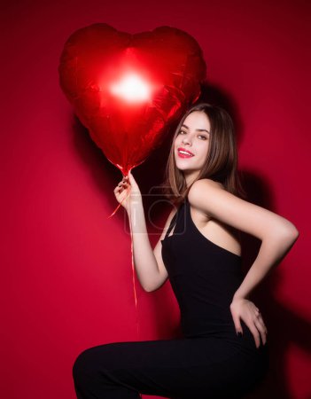 Foto de Hermosa joven en vestido negro sosteniendo globos corazón rojo, día de San Valentín. Cita romántica de mujer sexy. Mujer celebrando el Día de San Valentín. Retrato mujer bonita con globo de corazón rojo - Imagen libre de derechos