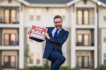 Photo for Funny realtor agent is a realtor with sign for sale in hand against the background on new apartment home background. Realtor in suit, outdoor portrait. Realtor offering new home. Property concept - Royalty Free Image
