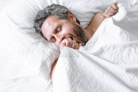Photo for Male head on pillow. Gray hair man yawning in white bed. Senior man yawning in bed at bedroom. Mature man yawning at home at morning. Good sleep. Millennial man yawningin bed - Royalty Free Image