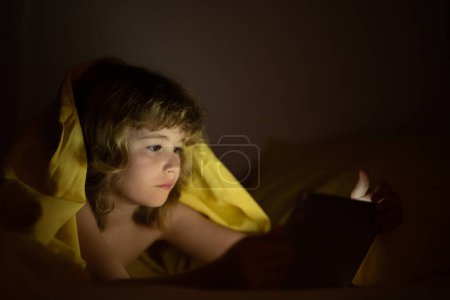 Téléchargez les photos : Il joue sur une tablette numérique. Enfant utilisant une tablette pour jouer et apprendre en ligne tout en étant allongé sur le lit, réflexe de lumière écran sur son visage. Tablette de jeux pour enfants sous couverture la nuit dans une pièce sombre - en image libre de droit