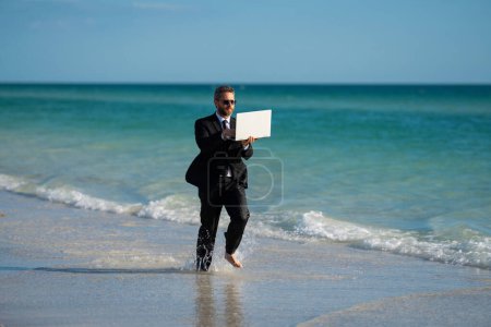 Photo for Businessman tourist in casual suit barefoot walking on tropical beach with laptop. Handsome business man on summer vacation. Businessman running on sea holding laptop. Businessman jumping in sea - Royalty Free Image