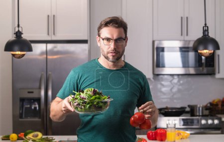 Téléchargez les photos : Homme avec une assiette de salade de légumes dans la cuisine. Homme cuisine salade végétalienne saine dans la cuisine. Homme millénaire à la cuisine moderne avec des légumes, préparer une salade de légumes frais pour le dîner ou le déjeuner - en image libre de droit