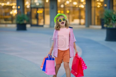 Foto de Increíble niño feliz emocionado con paquetes de compras en las manos. Comprador con bolsa de compras al aire libre. Venta de verano - Imagen libre de derechos