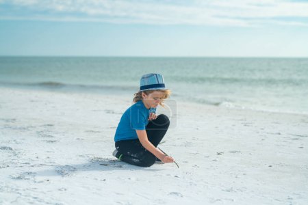 Téléchargez les photos : Garçon dessinant sur le sable au bord de la mer. Enfant dessinant du sable par imaginaire sur la plage pour apprendre. Enfant drôle jouant en plein air sur la plage d'été. Enfant émotionnel surpris en vacances d'été. Vacances d'été - en image libre de droit