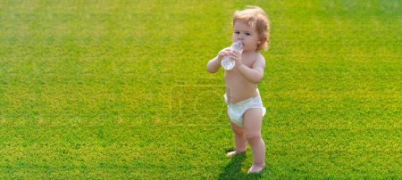 Photo for Baby on spring grass field, banner. Baby drinking water outdoor. Little child walking barefoot on green grass in sunny summer evening. Healthy child - Royalty Free Image