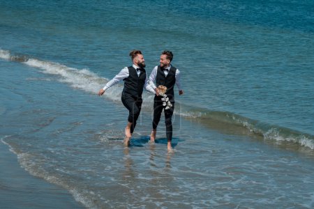 Foto de Hombre gay con pareja el día de la boda. Los novios gay caminando juntos en la playa del mar durante el día de la boda. Hombres románticos en el agua de mar. Retrato de feliz pareja gay en su boda al aire libre - Imagen libre de derechos