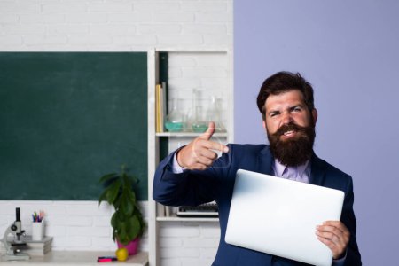 Photo for Modern male teacher holding laptop, waiting for students, smiling confidently. Teacher with notebook in classroom at school. Young male teacher with pointing finger near blackboard in classroom - Royalty Free Image