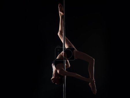 Foto de Chica flexible hace ejercicios gimnásticos en el pilón para estirar. Hermosa mujer flexible sexy demuestra cuerpo flexible sexy. Flexibilidad estiramiento en negro. Cuerpo femenino flexible. Baile poste - Imagen libre de derechos