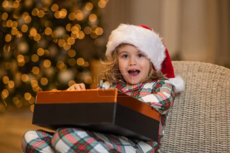 Foto de Feliz chico sorpresa abrir un regalo de Navidad. El niño sostiene una caja en un embalaje festivo. El muchacho se alegra en la fiesta, la Navidad, el Año Nuevo. El chico se levanta contra el árbol de Navidad - Imagen libre de derechos