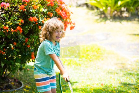Foto de Pequeño jardinero niño ayudando a regar flores con manguera de jardín en el jardín de verano. Trabajos de jardinería estacional - Imagen libre de derechos