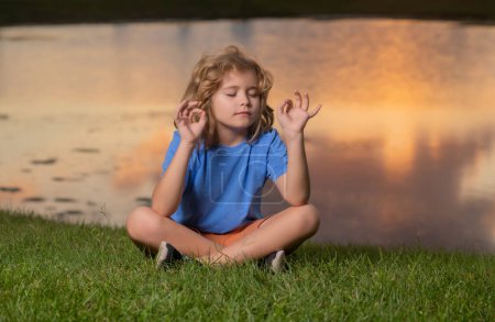 Téléchargez les photos : Enfant méditant en position lotus sur l'herbe, relaxant sur la nature estivale. Petit enfant heureux médite sur l'herbe verte le jour de l'été - en image libre de droit
