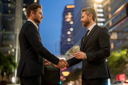 Photo for Businessmen shaking hands and giving money outdoor at nigth city street. Receiving money. Corrupted businessman giving away dollar money - Royalty Free Image