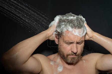 Foto de Hombre de mediana edad lavando el cabello en el baño. Cabezal de ducha en la bañera. Cara en espuma en la ducha. Bañista duchándose. Un tipo cercano duchándose. Concepto ducha. El hombre está bajo las gotas de agua en las duchas - Imagen libre de derechos