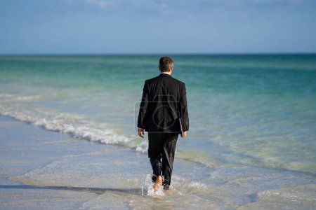 Foto de Vista trasera del hombre de negocios espalda en traje en agua de mar en la playa. Locas vacaciones de negocios. Hombre de negocios divertido con portátil en ropa formal cerca de la playa tropical. Caliente verano de negocios - Imagen libre de derechos
