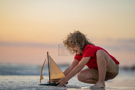 Photo for Little kid boy play with toy boat in the sea waves at the beach during summer vacation. Childhood and summer family vacation. Dream on travel. Travelling tour on sailing ship. Kids dream concept - Royalty Free Image