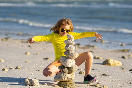 Photo for Excited kid boy playing with stones on the beach. Child play with pyramid of stones on the beach, sea seascape, rest and seaside kids summer vacation. Child playing on beach and building stone tower - Royalty Free Image
