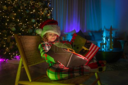 Foto de Niño en sombrero de santa en frente de fondo árbol de Navidad noche casa en Nochebuena. Lindo niño abriendo caja de regalo en la habitación decorada para la noche Navidad - Imagen libre de derechos