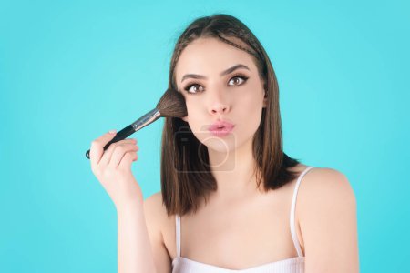Téléchargez les photos : Young woman applies powder on the face using makeup brush. Beautiful girl doing contouring apply blush on cheeks. Face beauty cosmetics. Fresh skin and natural make up. Powder blush on facial skin - en image libre de droit