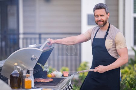 Foto de Hombre guapo de los 40 preparando la barbacoa. Cocine carne de cocina masculina en la parrilla barbacoa. Chico cocinando carne en la barbacoa para la cena familiar de verano en el patio trasero de la casa - Imagen libre de derechos