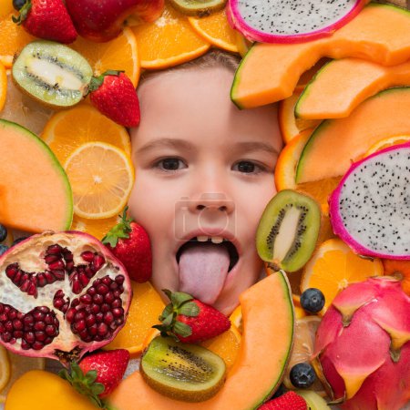 Téléchargez les photos : Un gamin lèche la fraise. Des fruits drôles. Visage enfant avec mélange de fraise, myrtille, fraise, kiwi, fruit du dragon, grenade, orange et melon. Mélange assorti de fruits près du visage de l'enfant - en image libre de droit
