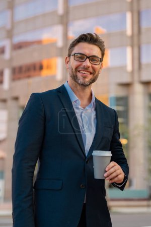 Foto de Hombre de negocios alegre en vasos con café para ir caminando por la calle hacia el moderno edificio de oficinas. Hombre de negocios hispano sonriendo feliz en la ciudad. Retrato de hombre de negocios al aire libre - Imagen libre de derechos
