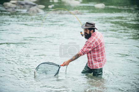 Photo for Young man fishing. Fisherman with rod, spinning reel on river bank. Man catching fish, pulling rod while fishing on lake. Wild nature. Catching trout fish in net - Royalty Free Image