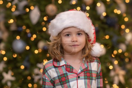 Téléchargez les photos : Joyeux enfant drôle dans le chapeau de Père Noël près de l'arbre de Noël. Noel et Nouvel An concept - en image libre de droit