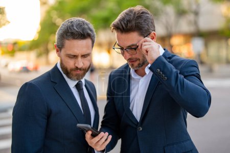 Foto de Dos hombres de negocios que usan mensajes de texto de aplicaciones de teléfonos móviles fuera de la oficina en la ciudad urbana. Empresarios sosteniendo teléfono inteligente para el trabajo de negocios. Exitoso hombre de negocios escribiendo en el teléfono inteligente al aire libre - Imagen libre de derechos