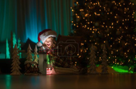 Téléchargez les photos : Cadeaux d'ouverture d'enfant la veille de Noël. Enfant en pyjama de Noël profitant soirée de vacances d'hiver à la maison près de l'arbre de Noël de nuit - en image libre de droit