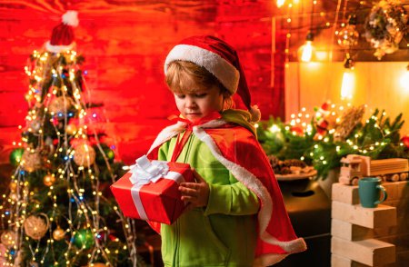 Foto de Feliz niño junto al árbol de Navidad mirando su regalo de Navidad. Niño pequeño lleva ropa de Santa Claus - Imagen libre de derechos