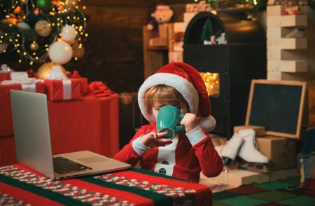 Téléchargez les photos : Le petit enfant porte des vêtements de Père Noël assis près de son ordinateur portable qui boit une boisson chaude de Noël. Fond de cheminée. Noël. Petit garçon. Père Noël - en image libre de droit