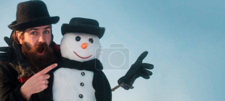Foto de Feliz Navidad y feliz año nuevo. Hombre de nieve de invierno divertido en el juego de sombreros con muñeco de nieve al aire libre. Hombre apuntando sobre muñeco de nieve en invierno fondo al aire libre. Hombre de nieve para bandera de invierno - Imagen libre de derechos