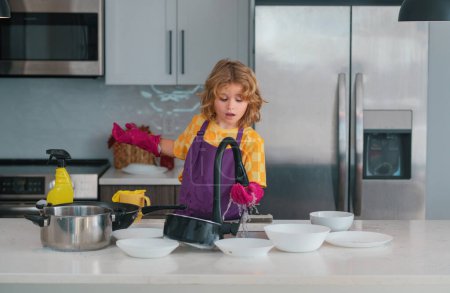 Téléchargez les photos : Nettoyage à la maison. Il essuie la vaisselle dans la cuisine. Nettoyage des enfants et ménage à la maison. Garçon mignon aider à l'entretien ménager dans la cuisine, nettoyage - en image libre de droit