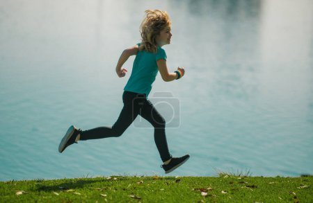 Téléchargez les photos : Enfants courir ou faire du jogging près du lac sur l'herbe dans le parc. Enfant sportif garçon coureur courir dans le parc d'été. Des enfants actifs, des enfants sportifs. Course matinale avec enfants - en image libre de droit