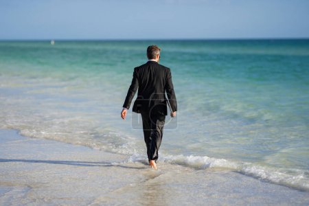 Foto de Trabajo independiente, negocio en línea. Vista trasera del hombre de negocios espalda en traje en agua de mar en la playa. Hombre de negocios divertido en un traje de negocios con portátil de pie en el agua. Trabajo remoto. Freelancer loco - Imagen libre de derechos