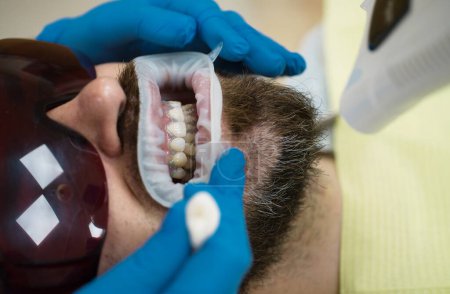 Téléchargez les photos : Jeune homme souriant après visite dentiste sur fond flou lumineux. Joli jeune homme souriant regardant caméra. Dentiste examinant les dents des patients en clinique - en image libre de droit