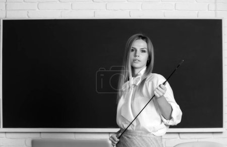 Foto de Joven profesor serio apuntando a la lección. Linda mujer joven con puntero enseñanza cerca de pizarra - Imagen libre de derechos