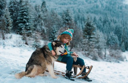 Foto de Niños felices divirtiéndose y montando en el trineo en el bosque nevado de invierno, disfrutar de la temporada de invierno. Niños abrazo abrazo perro husky - Imagen libre de derechos