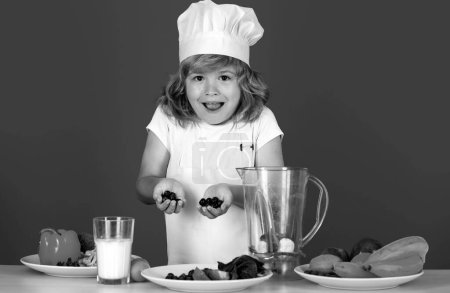 Téléchargez les photos : Chef cuisinier excité. Enfant chef habillé tablier de boulanger et chapeau de chef cuisine smoothie aux myrtilles isolé sur fond de studio. Alimentation saine pour enfants - en image libre de droit