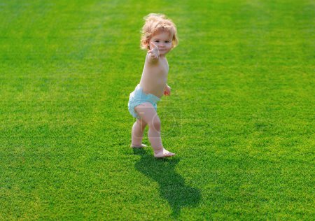 Photo for Baby walking barefoot on green grass in sunny summer evening. Little boy playing in the summer park in diaper pants - Royalty Free Image