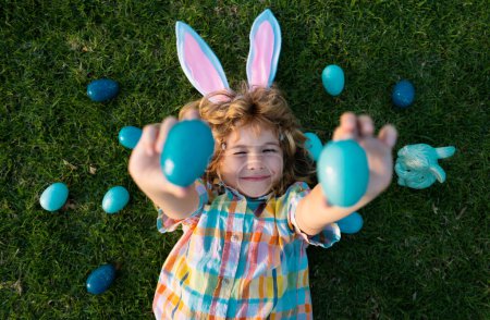 Foto de Conejito de Pascua niño cazando huevos al aire libre Feliz día de Pascua, Niño que pone en la hierba en el parque. Top view niños divertidos que ponen en la hierba - Imagen libre de derechos