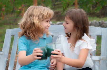 Foto de Two children drink smoothie, kids summer cocktail outdoors. Portrait of adorable brother and sister smile and laugh together while sitting on swing outdoors. Happy lifestyle kids - Imagen libre de derechos