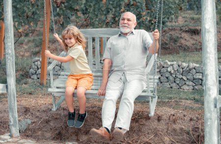Téléchargez les photos : Enfant garçon et grand-père balançant dans le jardin d'été. Grand-père et petit-fils assis sur balançoire dans le parc. Générations âges - en image libre de droit