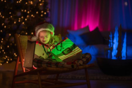 Téléchargez les photos : Cadeau cadeau ouvert enfant avec lumière magique. Éclairage cadeau sac cadeau. Enfant garçon 7-8 ans en pyjama de Noël profitant d'une soirée de vacances d'hiver à la maison près de l'arbre de Noël de nuit - en image libre de droit