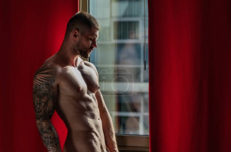Téléchargez les photos : Man standing near window. Seductive gay. Muscular body of man in hotel room. Sexy guy with naked body. Nude muscular man body with tattoo. Sexy man with muscular chest in bedroom on window curtains - en image libre de droit