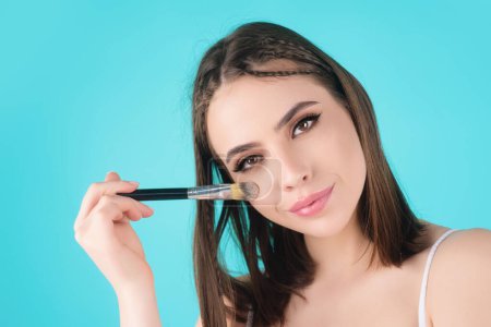 Téléchargez les photos : Studio portrait of a woman applying cosmetic tonal foundation on face using makeup brush. Beautiful girl doing contouring apply blush on cheeks isolated on studio background - en image libre de droit