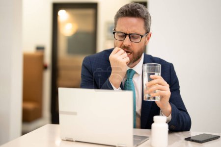 Photo for Man taking a medicine pill from headache migraine. Man in suit uses a laptop, is tired got headache migraine. Headache pain concept. Painful men are stressed at work in office. Stressful and headache - Royalty Free Image