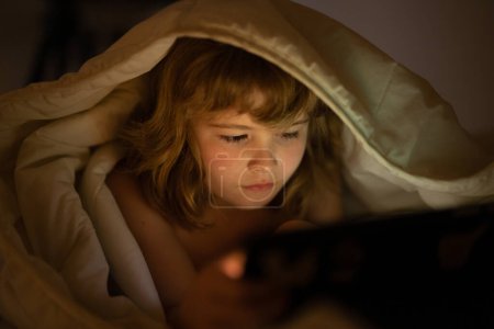 Photo for Kid playing game on tablet in bed at night. Kids with social media. Child lying in bed playing a tablet in dark room, light under blacket. Close up of kid watching cartoons on tablet - Royalty Free Image