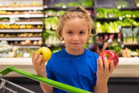 Foto de Niño con manzana y carrito de compras en el supermercado. Compras en el supermercado. Niños comprando comestibles en el supermercado. El niño compra verduras frescas en la tienda de comestibles. Niño en la tienda - Imagen libre de derechos