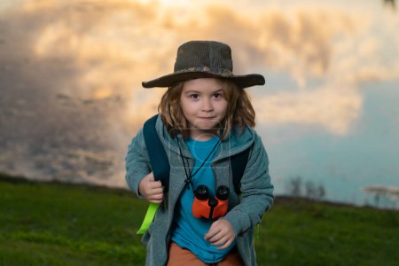 Foto de Un turista viajero. Lindo niño rubio con prismáticos con sombrero explorador y mochila en la naturaleza. Niño explorador senderismo y aventura - Imagen libre de derechos